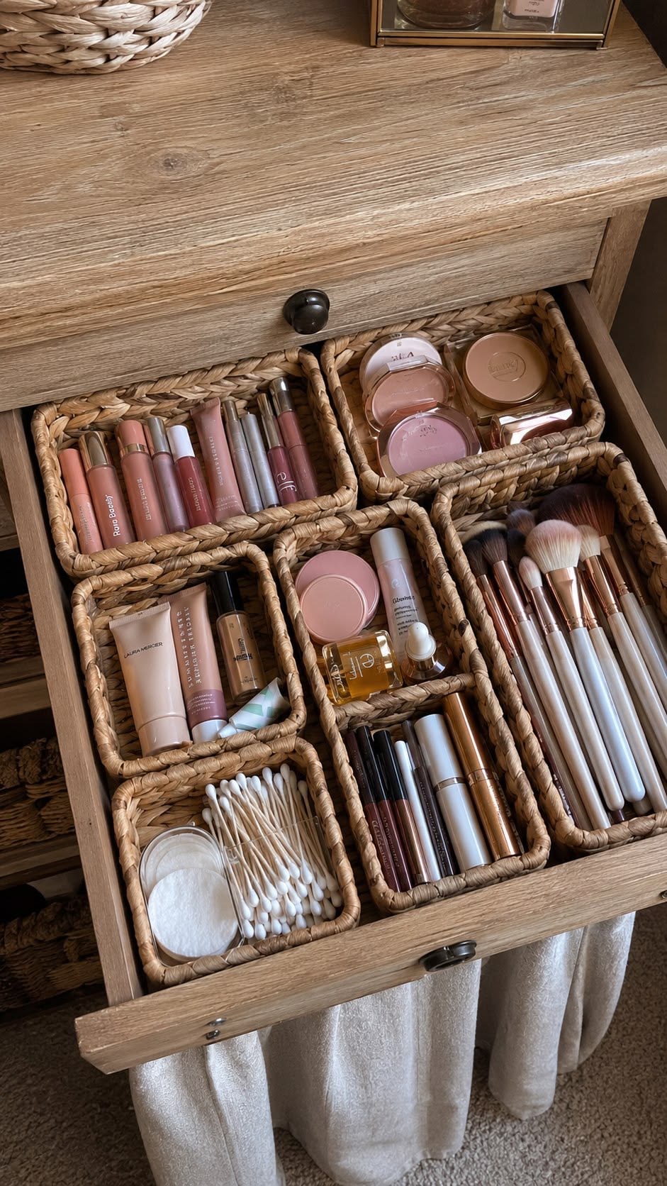 A wooden dressing-table drawer organised with woven baskets holding makeup and brushes